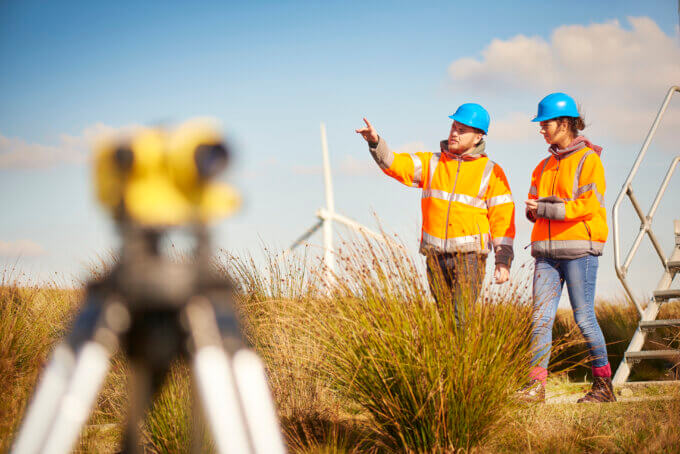 two wind farm engineers using a builder's level to plan out the expansion of the wind farm site. they are wearing orange hi vis jackets and blue hard hats . one is male , one is female. In the foreground the female is looking through the level whilst the male engineer is approaching .In the background wind turbines can be seen across the landscape. 