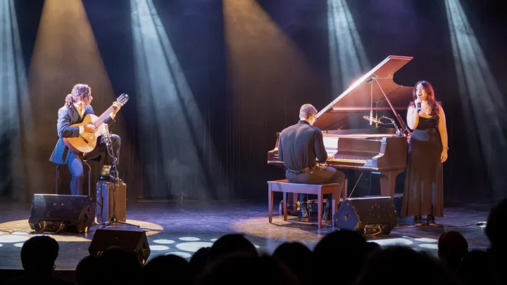 Trois étudiants performent sur scène lors de Cégeps en spectacle : un étudiant joue de la guitare acoustique, un autre est au piano à queue et une étudiante chante, un micro à la main.
