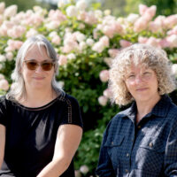 Deux femmes sont assises côte à côte devant un massif de fleurs, la photo coupée jusqu'à la taille et dans une ambiance ensoleillée.