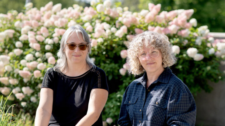 Deux femmes sont assises côte à côte devant un massif de fleurs, la photo coupée jusqu'à la taille et dans une ambiance ensoleillée.