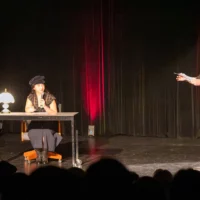 Sur la scène d'une salle de spectacle, deux personnes étudiantes se font face. Une étudiante est assise à une table recouverte d'une lampe, un micro et un stylo dans les mains. Elle regarde son partenaire de chant, qui lui parle debout dans un micro. La silhouette du public est visible au premier plan, devant la scène.