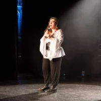 Une étudiante vêtue dans un style médiéval, avec une tunique blanche à manches larges recouverte d'un corset, chante debout sur une scène de salle de spectacles, éclairée par un projecteur.