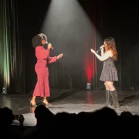 Sur la scène d'une salle de spectacles, derrière la silhouette d'un public visible au premier plan, deux étudiantes se font face, un micro à la main et chantent en se regardant.