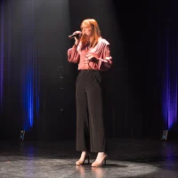 Une étudiante, un micro à la main, chante debout sur la scène d'une salle de spectacles, éclairée par des projecteurs.