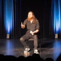 Un étudiant assis sur une chaise au centre de la scène d'une salle de spectacles s'adresse au public dont on devine la silhouette au premier plan.