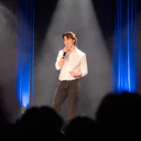 Un étudiant, les yeux fermés, chante sur la scène d'une salle de spectacles, éclairée par une lumière de projecteur. La silhouette du public est visible au premier plan.