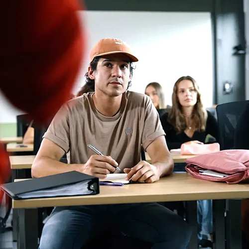 Un élève écoute dans une classe assis dans une salle avec d'autres élèves en arrière de lui et la silhouette de la personne enseignante sur l'avant.