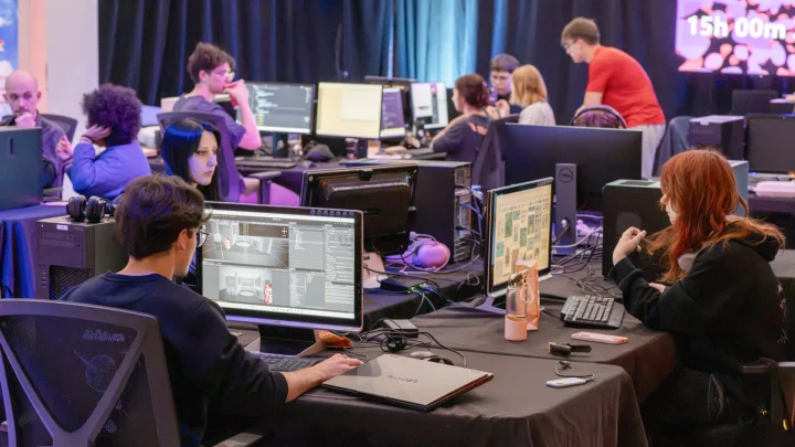 Des étudiantes et étudiants participent au Webjam, un marathon de 24 heures de création numérique : on voit le décompte du chronomètre sur un écran au loin et des élèves qui travaillent sur leur poste de travail respectif, dont un étudiant au premier plan qui fait de la programmation de code.